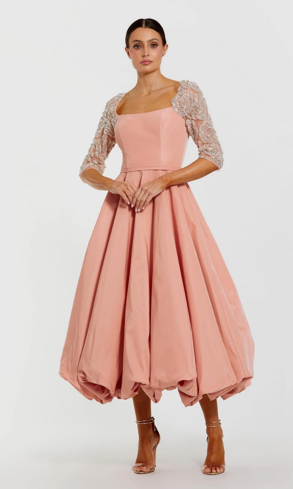 A woman models the Mac Duggal 2320 Semi-Formal Midi Party Dress in rose, featuring a square neckline, embellished mesh sleeves, bubble hem, and paired with strappy heels for an elegant look. Her hair is tied back.